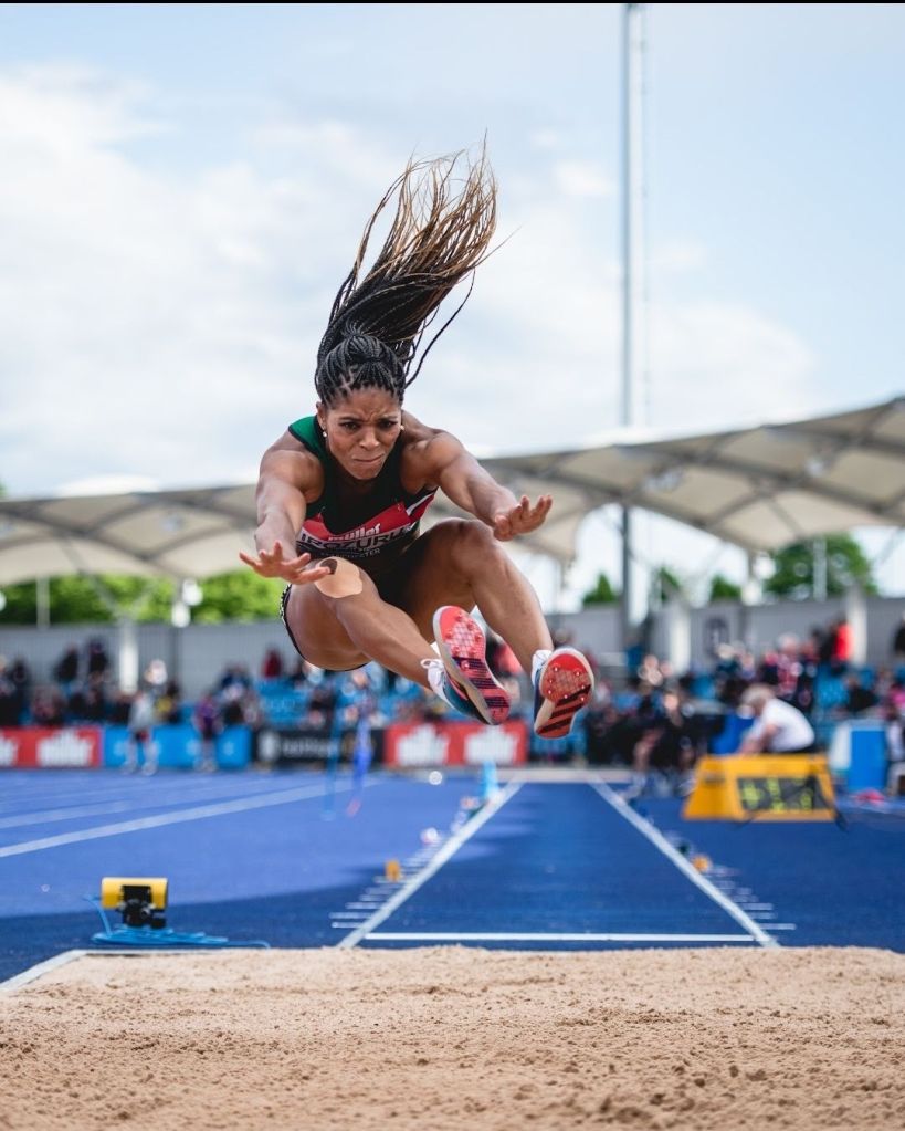 Abigail - a black woman - jumping through the air - arms reaching forward towards feet to land in the sand pit.